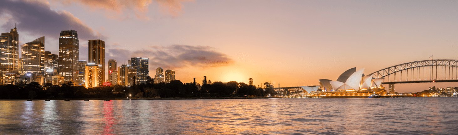 Twilight over a waterfront cityscape, featuring the iconic opera house and a distinctive bridge, with reflections on the water.