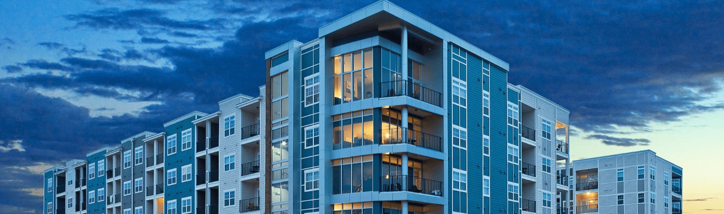 Captivating image of a modern apartment building in the United States at dusk, with its sleek, glass facade reflecting the stunning hues of the sunset sky. The building's angular corner provides a unique perspective, highlighting the blend of contemporary architecture and the natural beauty of the evening sky.